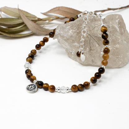 A beaded bracelet with brown and black tiger's eye beads, silver spacer beads, and a silver charm, displayed on a white surface next to a clear crystal and dried leaves.