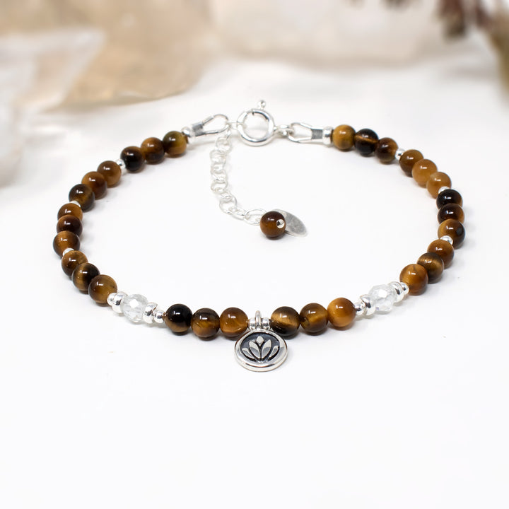 A beaded bracelet with brown and black tiger's eye beads, clear faceted beads, and sterling silver accents, including a small lotus flower charm, displayed on a white surface.